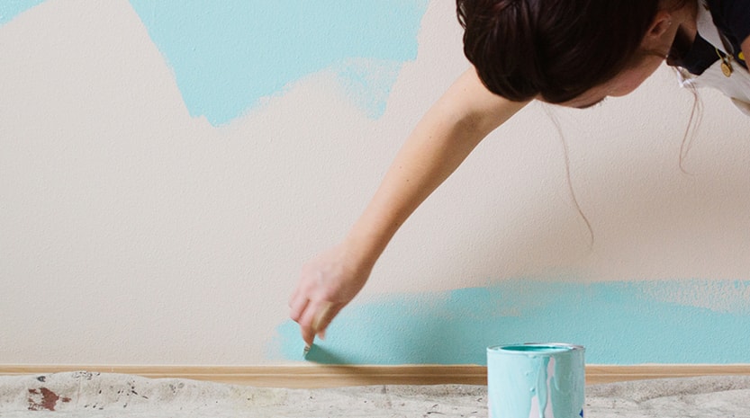 A woman with her hair tied back leans down to continue painting the bottom of her walls blue. Drop cloth is covering the floor and a paint can sits on the floor under her.