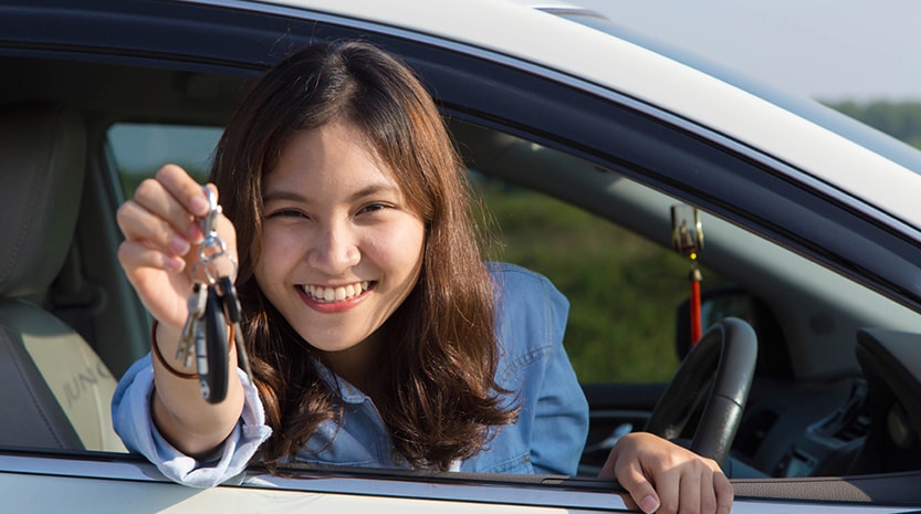 A young woman leans excitedly out of her car window on a summer day dangling her keys close to the camera.
