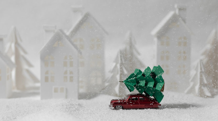 A toy car carrying a toy Christmas tree through a fake snowy landscape.
