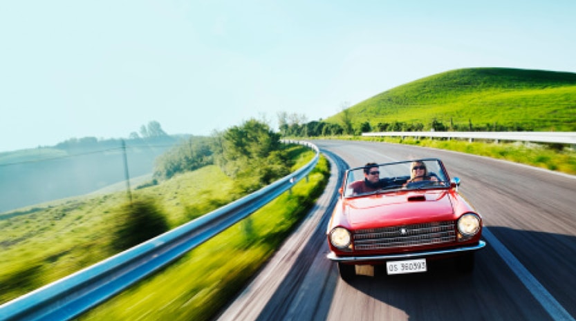 A man and a woman speed down a countryside road in an old red convertible. The road and surrounding grass is blurry to indicate speed.