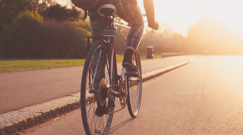 A person wearing a backpack rides their bicycle down a paved path into the sunset.