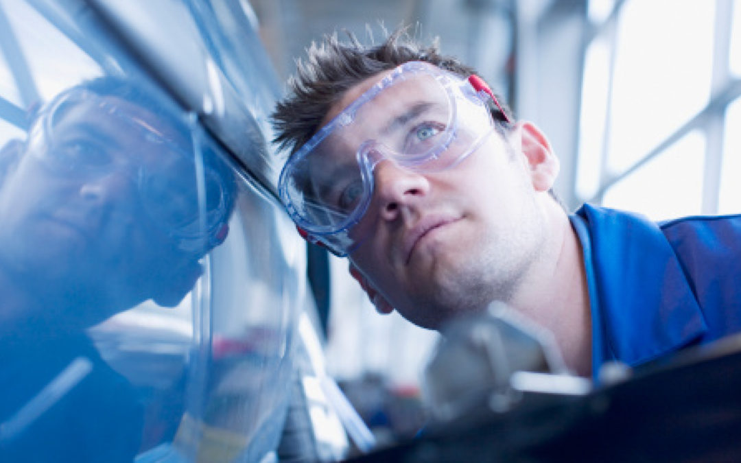 A man wearing safety goggles holds a clipboard and leans down close to the side of a car.