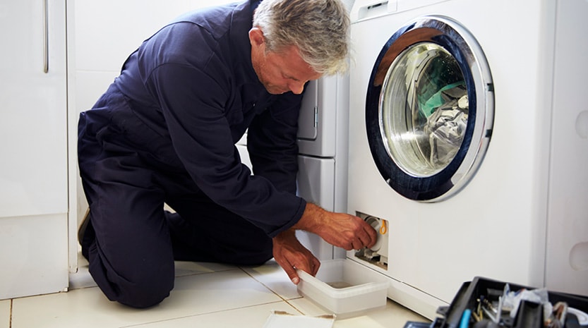 A worker leans over on his knees to fix a washing machine.
