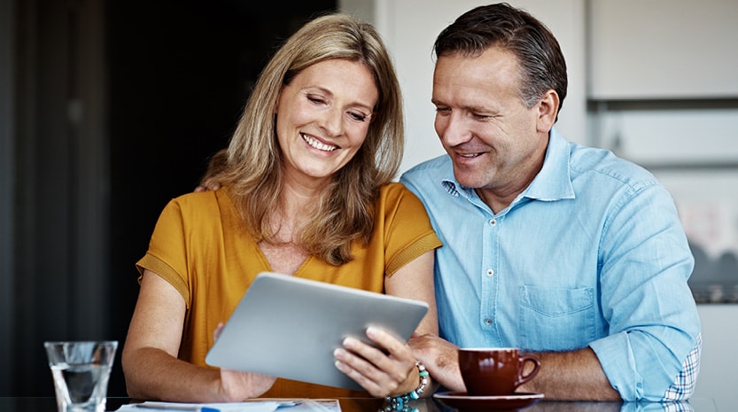 An older couple smiles while looking at a tablet at the kitchen table.