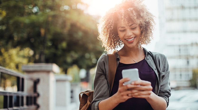 A young woman with a bag over her shoulder looking at her phone on a sunny sidewalk.