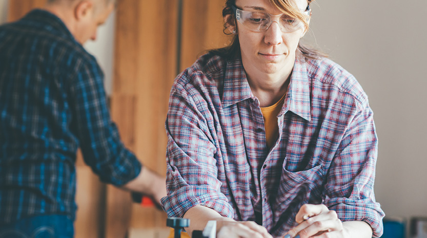 A woman wearing flannel and safety glasses leans over a work bench with a pencil in her hand. There is a man doing other things in the background.