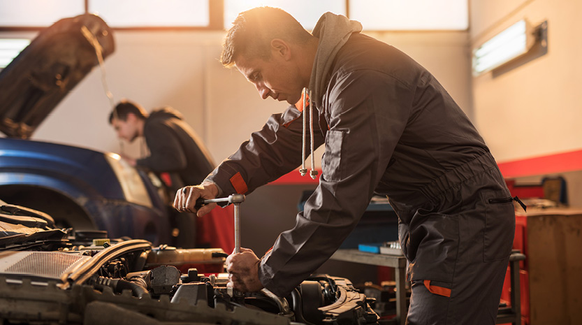 A mechanic leans over to fix something in the open hood of a car in an auto repair shop.