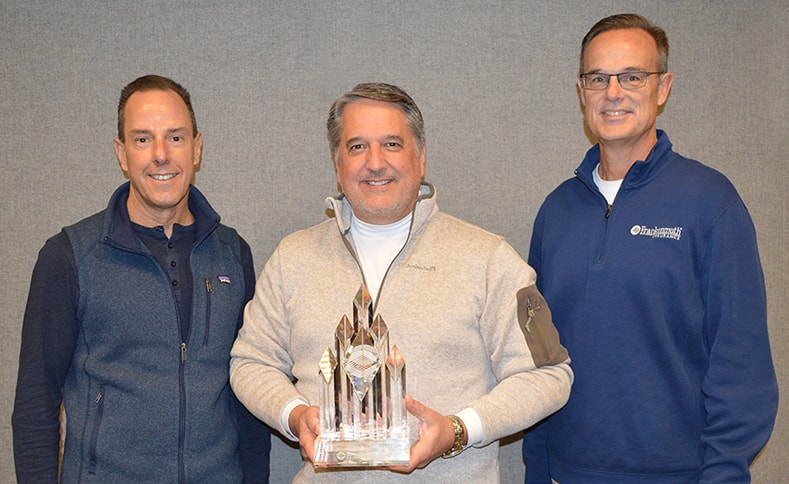 Three men stand against a gray wall, smiling for the camera. The man in the middle is holding a geometric glass award trophy.